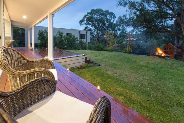 A covered outdoor deck or veranda with timber decking, white structural posts and ceiling, and wicker lounge chairs in the foreground. The deck overlooks a broad grassy backyard with mature trees, a small timber structure, and an outdoor fire pit, creating a relaxed evening entertaining setting.