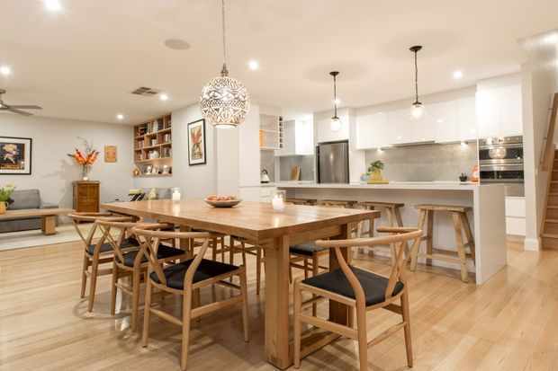Open-plan kitchen and dining area with a large timber dining table, matching wooden chairs, pale timber flooring, white cabinetry, a tiled kitchen splashback, pendant lighting, and built-in shelving. A living area is visible to the left, creating a warm, contemporary family space.