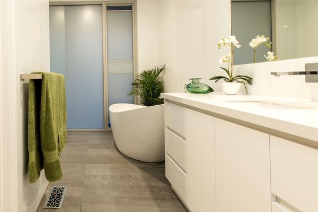 Contemporary bathroom with a freestanding white bathtub, large white vanity, integrated basin, wall mirror, frosted-glass door, grey tiled floor, and green towel and plants adding natural colour.