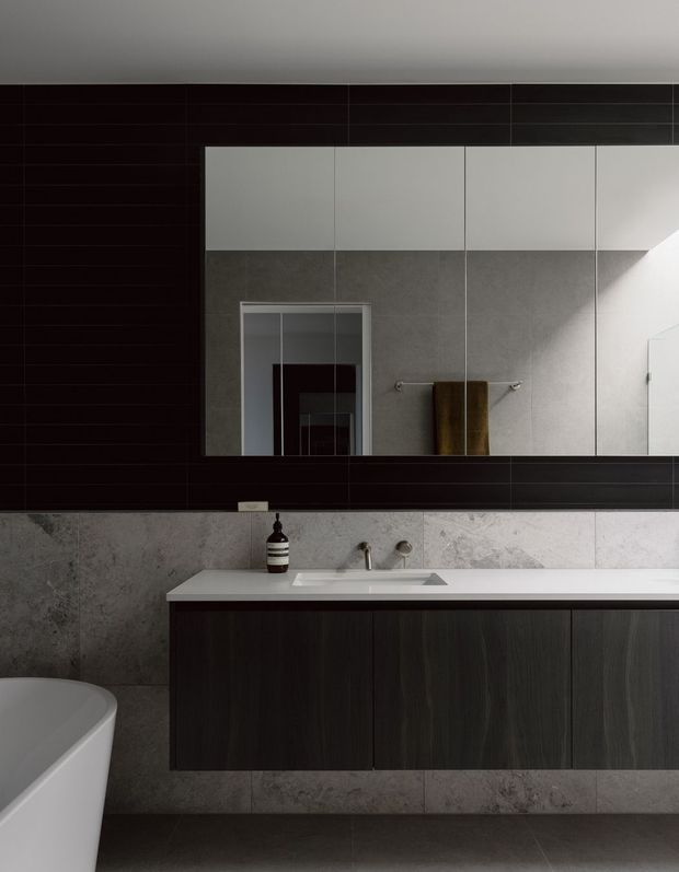 Contemporary bathroom vanity with a long dark wood-look floating cabinet, white integrated basin and benchtop, grey stone-look tiled walls, and a large mirrored cabinet above. A freestanding white bathtub is partially visible at left. Dark horizontal wall tiling and minimalist chrome fixtures create a moody, refined interior.