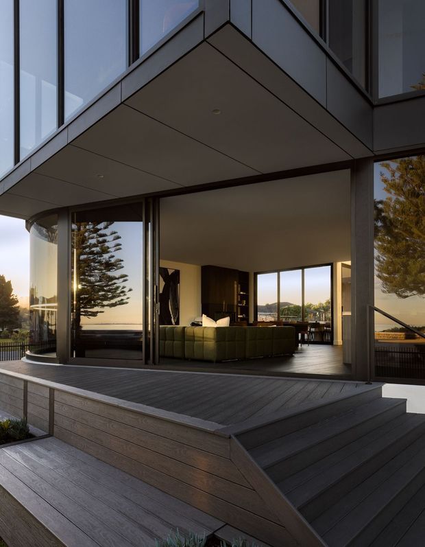 A contemporary multi-level home exterior viewed from a large timber deck and stepped outdoor terrace. The building has dark framed floor-to-ceiling glazing, projecting box-like upper volumes, and broad covered outdoor living areas. Through the open glazing, a living room and dining area are visible, with expansive views beyond. The image is captured in warm, low evening light and emphasizes the connection between indoor and outdoor spaces.