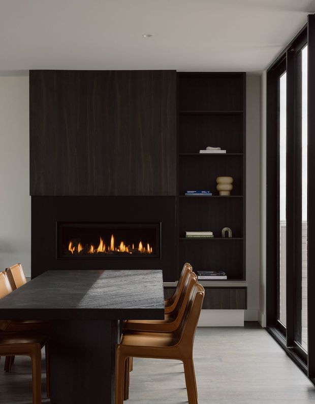 A contemporary dining area with a dark timber built-in feature wall incorporating a linear gas fireplace and open display shelving. A large dark stone dining table with brown leather dining chairs occupies the foreground, while tall black-framed glazing lines the right side. The space has a restrained, moody palette with warm firelight and abundant daylight.