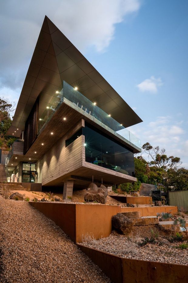 Exterior view of a dramatic modern multi-level house on a steep, landscaped site. The building features a sharply projecting angular roof canopy, extensive floor-to-ceiling glazing, dark timber wall cladding, timber soffits, exposed concrete supports, and glass balustrades. Terraced rust-toned retaining walls and gravel landscaping surround the elevated structure, with trees and sky in the background.