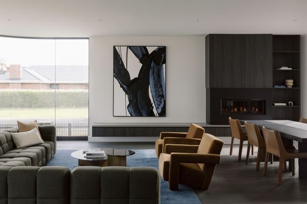 A refined open-plan living and dining room with a large glazed window, low-profile seating, sculptural lounge chairs, a dark built-in media and fireplace wall, and an integrated dining area. The room combines neutral plaster walls, dark timber joinery, blue-grey carpet, and warm brown upholstery. A large abstract artwork and linear recessed lighting add contemporary character.