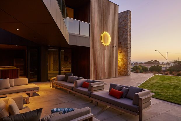 A sophisticated contemporary outdoor living terrace at dusk, adjoining a modern multi-level home. The space features several low-profile upholstered sofas and lounge chairs arranged around timber coffee tables, with warm integrated lighting beneath a deep overhang. The facade combines vertical timber slats, dark glazing, smooth dark panels, and a prominent natural stone-clad wall. A manicured lawn and coastal-looking horizon are visible beyond the terrace.