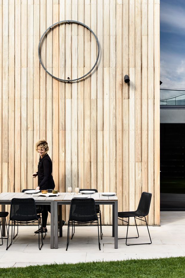 Contemporary outdoor dining area beside a modern building, featuring a long rectangular dining table with black woven chairs, pale timber vertical cladding, a large circular wall feature, and a dark glazed opening. The setting is bright and minimalist with a clean paved surface and lawn edge.