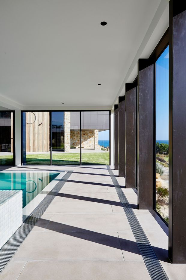 A bright, elongated indoor pool enclosure with full-height black-framed glazing along one side and at the far end. Large pale floor tiles are crossed by linear drainage grilles and strong geometric shadows. The turquoise swimming pool occupies the left foreground, while the glazing opens views toward a stone-and-timber exterior, lawn, blue ocean and coastal landscape. The space has a clean, contemporary architectural character with a white ceiling and recessed downlights.