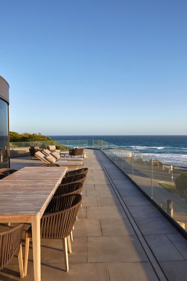 An expansive rooftop or upper-level oceanfront terrace overlooking the sea and beach. The paved terrace has a long timber dining table with dark woven chairs, several cushioned sun loungers, glass balustrades, and a curved glazed building edge at the left. The scene is sunlit and contemporary, with unobstructed coastal views.
