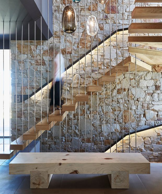 Interior architectural photograph focused on a sculptural floating staircase with slim dark metal balustrade cables, set against a prominently exposed irregular stone masonry wall. Warm timber stair treads and a substantial timber bench occupy the foreground, while pendant glass lights hang above. A blurred person is visible on the stairs, and daylight enters from glazing at left.