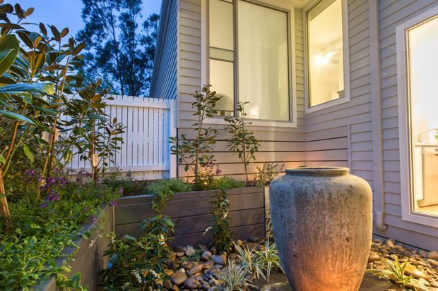 A warmly lit exterior courtyard or entry garden beside a contemporary house, featuring pale horizontal weatherboard cladding, white-framed windows, a white slatted boundary fence, raised timber garden beds, river stones, planting, and a large textured ceramic urn.