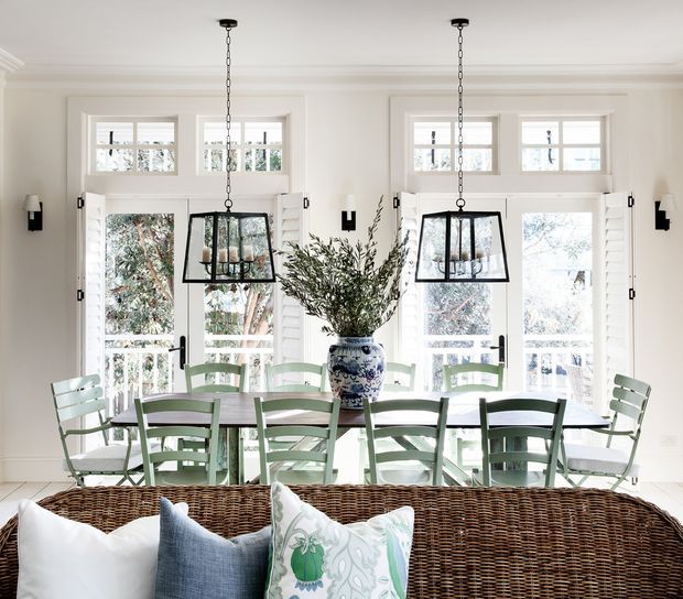 Bright, airy dining area with a long rectangular dining table surrounded by pale green chairs, positioned in front of white French doors and windows. Black pendant lanterns hang above the table, while a blue-and-white ceramic vase with leafy branches forms the centerpiece. A woven rattan sofa or bench with decorative cushions occupies the foreground. The space features white painted wall panelling, trim, and window-door frames with abundant natural light.