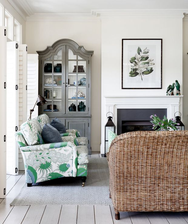 Bright, refined living room with a white-painted fireplace, traditional decorative mouldings, pale timber flooring, and a grey glass-front display cabinet. Patterned green-and-white armchairs sit beside a large woven rattan sofa, with botanical artwork and decorative accessories adding colour.