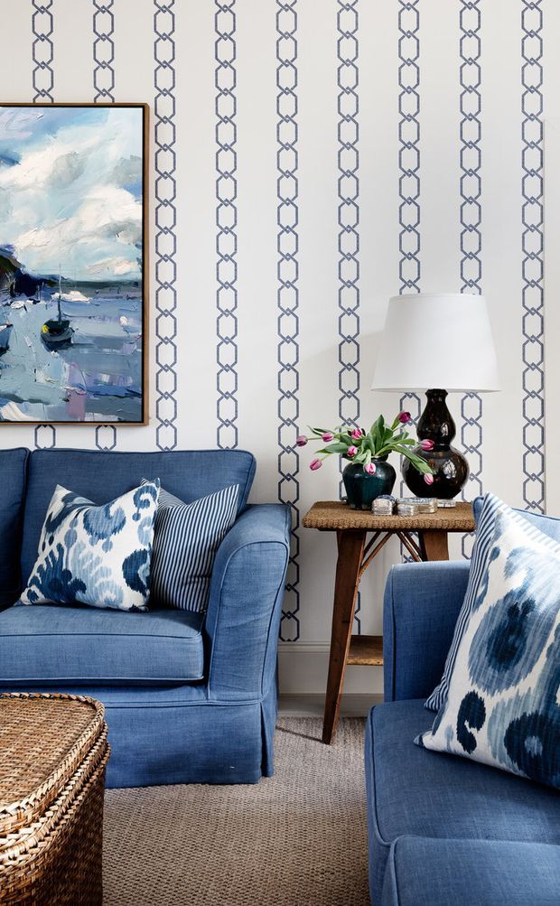 A styled living room featuring two deep blue upholstered sofas with patterned blue-and-white cushions, a woven coffee table, and a narrow timber side table with a lamp, dark ceramic vessels, and flowers. The walls are covered in a white and blue geometric trellis-patterned wallpaper, with a blue-toned abstract artwork framed in black. A neutral textured carpet provides contrast beneath the furniture.