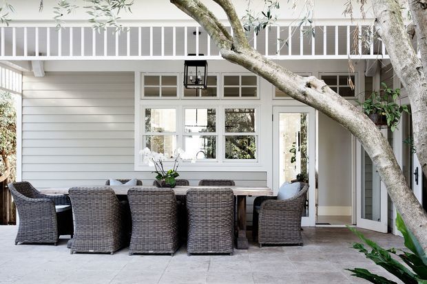 A bright covered outdoor dining terrace adjoining a light-painted residential facade. The space features a long timber dining table surrounded by woven outdoor chairs, pale tiled paving, horizontal wall cladding, multi-pane windows, a glazed door, and a white balustrade above. Mature tree branches frame the foreground.