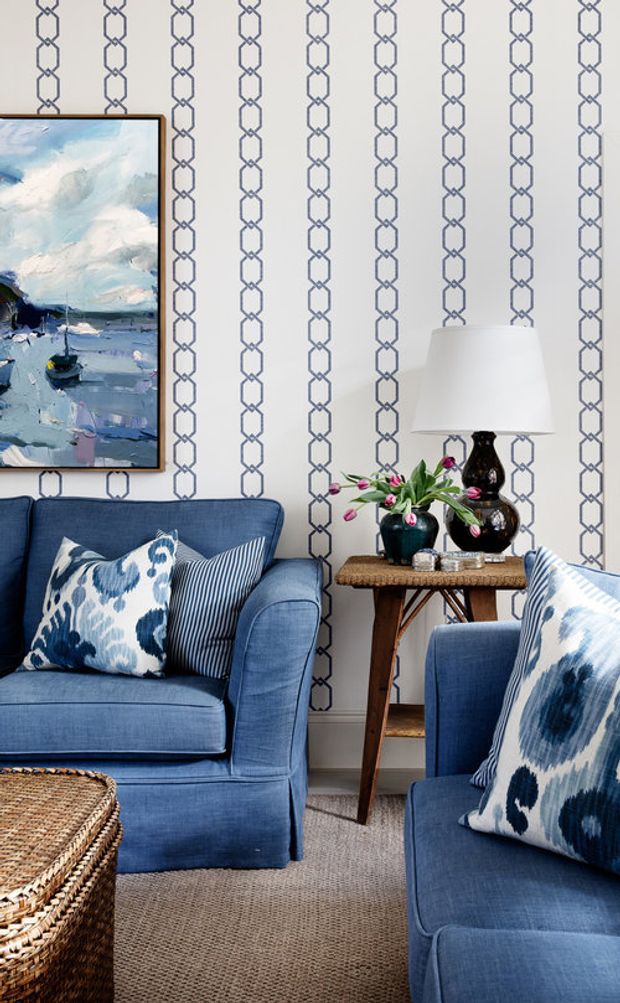 A styled living room corner featuring two deep blue upholstered armchairs and a matching sofa, patterned blue-and-white cushions, a woven coffee table, and a narrow wooden side table with a lamp, vases, and flowers. The room has striking white wallpaper with repeated navy geometric chain motifs, a blue-toned abstract artwork, and a neutral textured carpet.