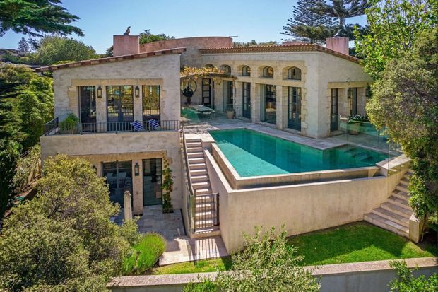 Exterior view of a large two-storey stucco residence with Mediterranean-inspired detailing, arched openings, clay-tiled roof edges, decorative cornices, balconies, terraces and a raised rectangular swimming pool surrounded by landscaped grounds.