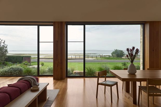 A warm, timber-lined open-plan living and dining space with full-height black-framed glazing overlooking a landscaped coastal setting and ocean beyond. The room features timber flooring, vertical timber wall cladding, a low upholstered lounge, and a simple timber dining table with chairs. Natural light and broad views create a calm, understated interior.