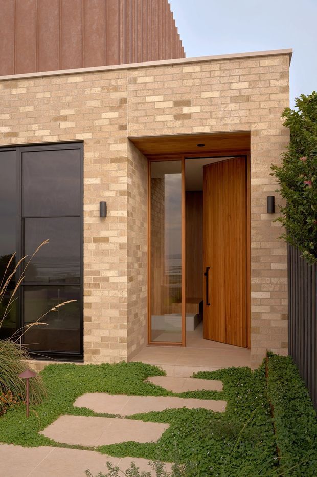 Contemporary residential entrance viewed from the landscaped front path. The facade combines pale variegated brickwork with a tall dark-framed window, a deeply recessed timber-lined entry, and a large warm timber pivot door. Stepping-stone paving runs through low groundcover planting, while vertical ribbed cladding rises above the brick volume.