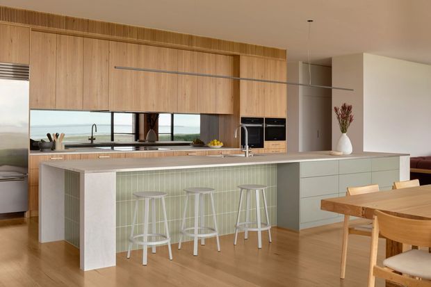 A bright open-plan kitchen and dining area with a large island featuring a pale stone benchtop, soft green vertically fluted tile facing, integrated pale cabinetry, and three white bar stools. Warm timber cabinetry extends to the ceiling with a slatted detail above, while a long horizontal window provides an expansive coastal view. A timber dining table and chairs sit in the foreground, and integrated dark ovens, a cooktop, sink, and pendant lighting complete the clean contemporary fit-out.