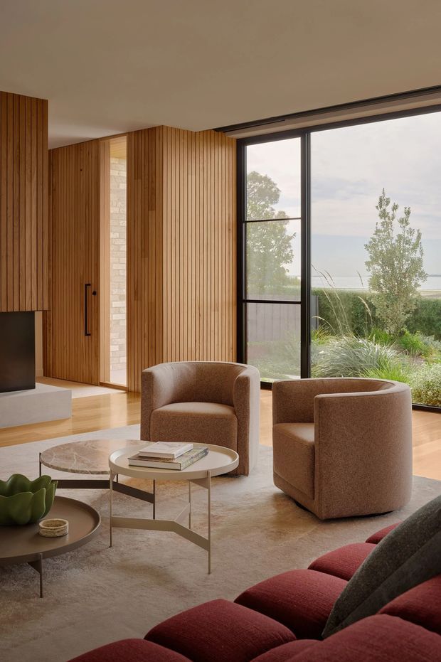 A warm, refined living room with floor-to-ceiling black-framed glazing overlooking a landscaped exterior. Vertical timber wall panelling and matching built-in joinery contrast with pale flooring and a light neutral rug. Two rounded upholstered lounge chairs sit around layered oval coffee tables, with a deep burgundy sectional sofa in the foreground. A pale stone or brick feature wall and concealed doorway add architectural detail.