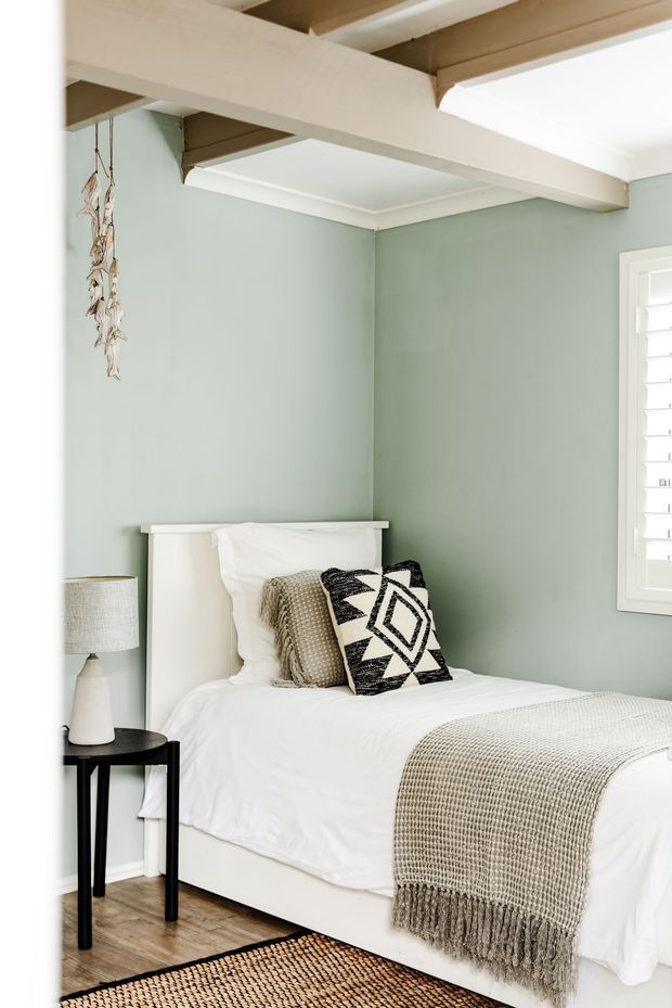 A bright, compact bedroom with soft sage-green walls, white bedding, a simple white headboard, and a textured neutral throw. A black round bedside table with a ceramic lamp sits beside the bed, while exposed painted ceiling beams and a white plantation-shutter window add architectural character. A patterned black-and-white cushion and woven rug provide graphic accents.