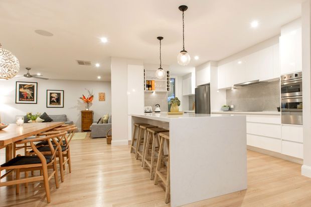 Bright open-plan kitchen and dining area with a large white island, integrated cabinetry, stainless-steel appliances, and a small patterned tile splashback. Timber bar stools, a timber dining table with matching chairs, light timber flooring, pendant lights, and contemporary artwork create a warm, inviting interior. A living area is visible beyond the dining space.