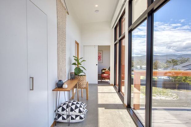 Bright contemporary hallway with full-height glazed windows and dark-framed sliding doors along one side, opening to a balcony and mountain landscape. The corridor has pale walls, a polished concrete floor, built-in white storage, a narrow timber console, indoor plants, patterned floor cushions, and a glimpse of a furnished room at the far end.