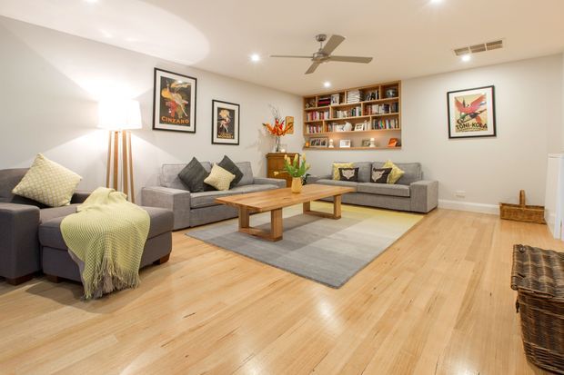 Bright, spacious living room with light timber flooring, grey sofas and armchairs, a large neutral area rug, a central timber coffee table, framed artwork, recessed lighting, a ceiling fan, and built-in timber shelving filled with books and decor.