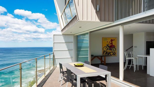 A bright contemporary coastal apartment terrace overlooking the ocean, with a large outdoor dining table, glass balustrade, sliding glazing, white vertical cladding, and an open-plan interior containing a dining area and kitchen. The architecture features strong horizontal and vertical lines, projecting upper-level massing, and extensive glazing.