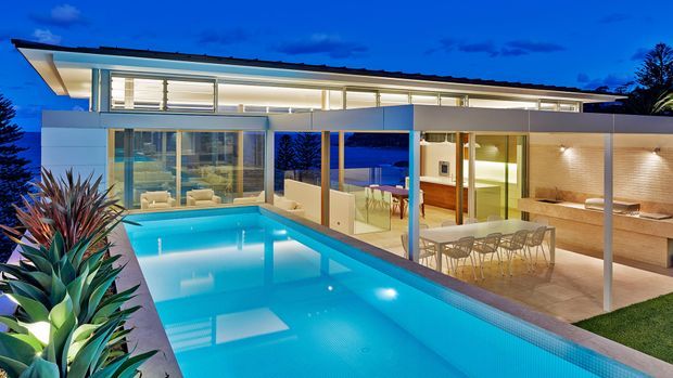 Twilight exterior view of a sleek modern multi-level residence organized around a large illuminated swimming pool. The home has extensive floor-to-ceiling glazing, a long low roofline, white rendered walls, timber-lined interior areas, and an open-plan dining and outdoor entertaining zone opening directly onto the pool terrace. Tropical architectural planting frames the foreground.