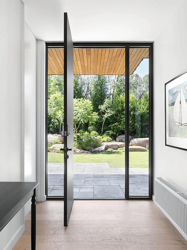 Bright minimalist interior entryway viewed toward a full-height glazed opening and garden beyond. A slim dark-framed glass door is open, with a timber-lined exterior soffit above the patio, pale timber flooring, white walls, a slim black console table, framed artwork, and a white panel radiator. The view includes a paved terrace and landscaped garden outside.