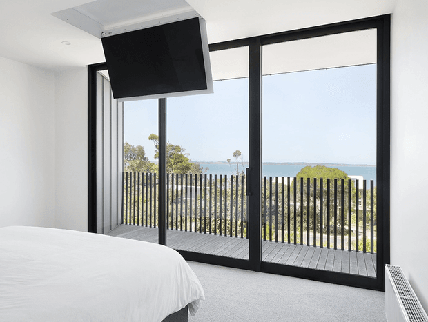 Bright modern bedroom with a partially visible white bed, pale carpet, and expansive black-framed floor-to-ceiling glazing opening onto a balcony with a vertical-slat balustrade and ocean view. A large black ceiling-mounted feature light or ventilation element hangs above the room.