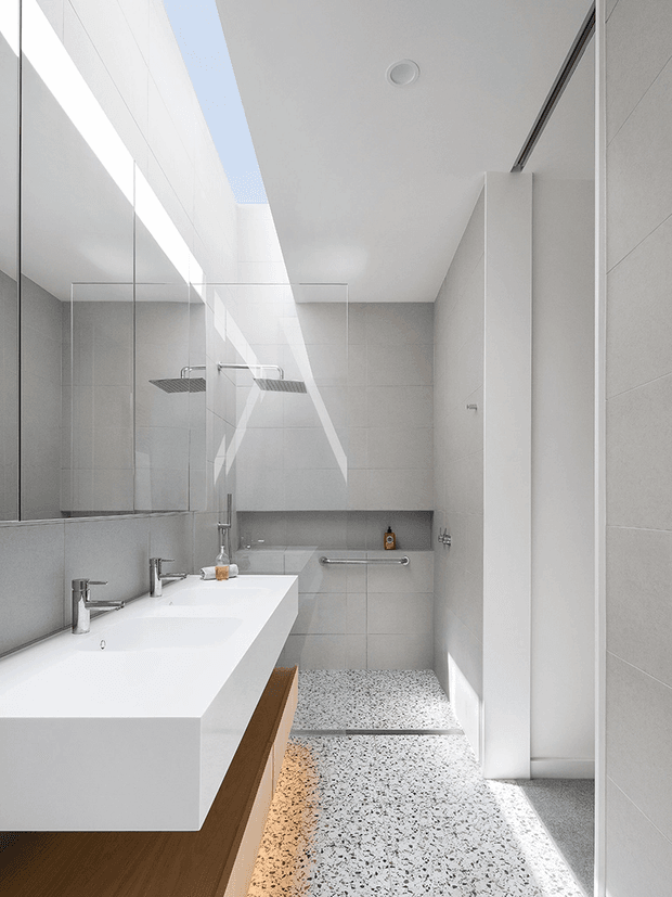 Bright contemporary bathroom with a long white double vanity, frameless mirror, wall-mounted chrome fixtures, a fully glazed walk-in shower, and a built-in bathtub at the rear. Large light-grey wall tiles and a terrazzo-style floor create a clean, minimalist finish, with strong natural light entering through a linear skylight or clerestory.