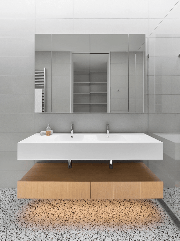 Minimalist contemporary bathroom featuring a floating double vanity with a white integrated basin and chrome wall-mounted taps, set above a timber vanity cabinet. Large grey wall tiles, mirrored storage cabinetry, and terrazzo-style flooring create a clean, restrained finish.
