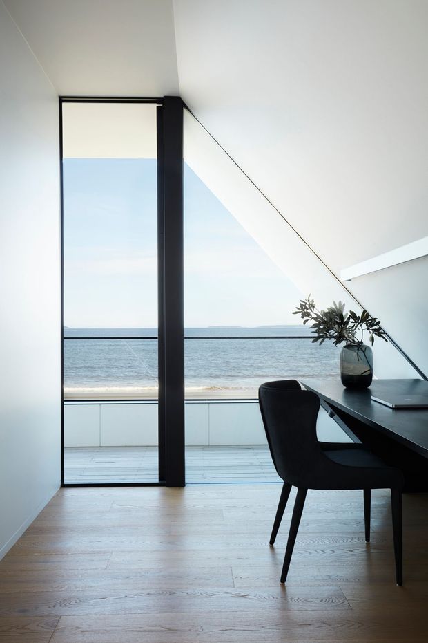 A minimalist home office or study overlooking the ocean through expansive floor-to-ceiling glazing. The room has white walls and ceiling, pale timber flooring, a black-framed glazed opening, and a dark built-in desk with a black upholstered chair. A simple dark vase with foliage sits on the desk, while a balcony or deck and beach are visible beyond the glazing. The composition is calm, bright, and highly pared back, with a strong coastal outlook.