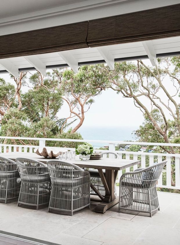 A bright covered outdoor dining terrace with a large timber dining table and woven outdoor armchairs. White railings, tiled flooring, exposed white pergola beams, and a dark woven roller blind frame views through coastal trees toward the blue sea. The setting feels relaxed, airy, and coastal.