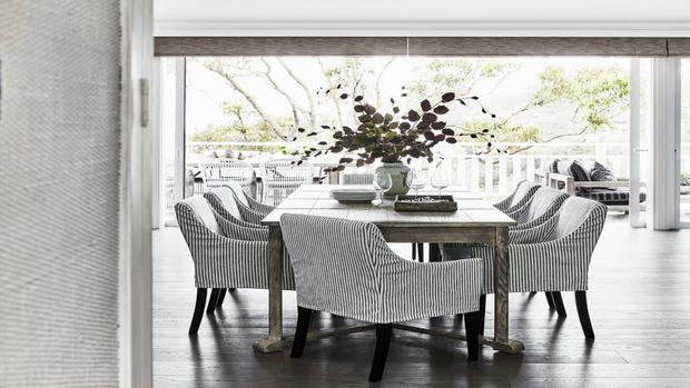 A bright dining room with a rectangular timber dining table surrounded by upholstered striped dining chairs. Large windows and doors open the room to a covered outdoor area, with pale walls, dark timber flooring, and a simple decorative branch arrangement on the table.