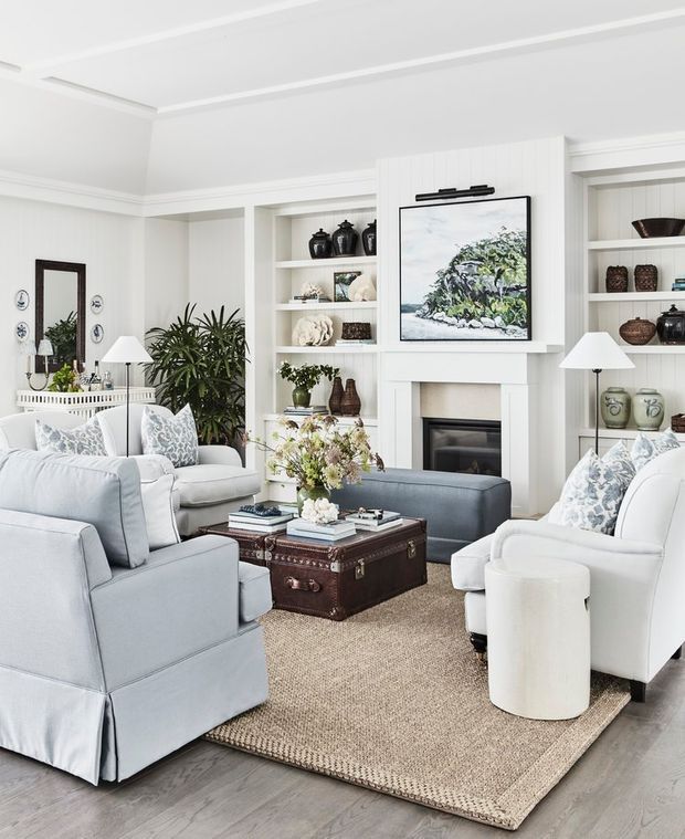 Bright, refined living room with a symmetrical seating arrangement around a leather trunk coffee table and fireplace. The room features pale upholstered sofas and armchairs, built-in white shelving filled with decorative objects, a white mantel, light timber flooring, a natural woven rug, botanical accents, and coastal-toned artwork and cushions.