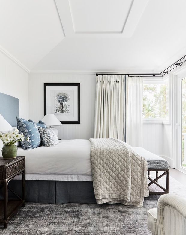 Bright, refined bedroom with a large upholstered bed, layered blue-and-white cushions, a quilted grey throw, dark bedside table, framed botanical artwork, white vertical wall panelling, and floor-to-ceiling curtains beside glazed doors. The room has a soft neutral palette with subtle coastal detailing and abundant natural light.