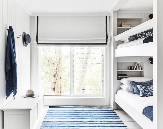 Bright, compact bunk room with built-in white bunk beds, integrated open shelving, navy-and-white patterned bedding, vertical white wall panelling, a large window with a simple Roman blind, and a striped blue-and-white rug. The room has a clean coastal-inspired feel and efficient built-in storage.