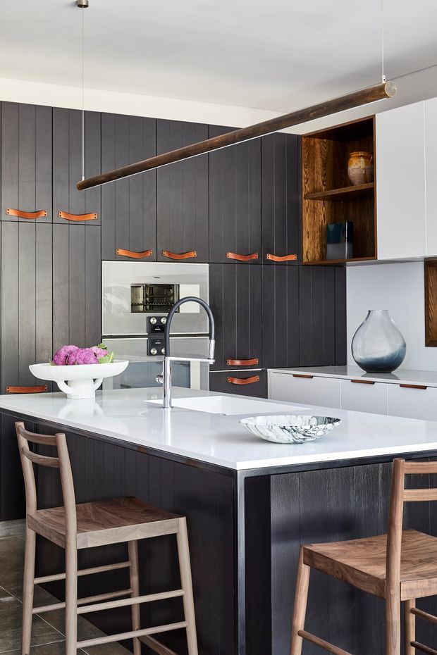 A refined contemporary kitchen centred on a large white island with an integrated sink and chrome gooseneck tap. The kitchen features dark vertically grooved cabinetry with warm leather drawer pulls, white lower cabinetry, a built-in oven, and an open timber display shelf. Timber bar stools sit at the island, while a long suspended timber pendant adds a strong linear feature overhead. Decorative ceramics and a floral arrangement provide subtle accents.