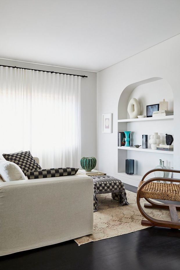A bright, refined living room with a large cream sofa, patterned black-and-white cushions and ottoman, a woven lounge chair, and a built-in white wall niche with curved arch detailing and display shelving. Sheer full-height curtains filter abundant daylight, while dark timber flooring and a neutral patterned rug provide contrast. Decorative ceramics, books, and framed artwork add restrained colour accents.