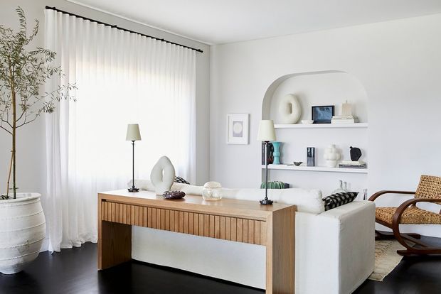 Bright, refined living room with a white sofa positioned behind a substantial timber console or desk, layered with sculptural neutral decor and table lamps. Full-height sheer curtains provide soft filtered light, while built-in curved shelving displays minimalist objects. The room has dark timber flooring, white walls, a large indoor plant, and a woven timber lounge chair, creating a calm contemporary interior.