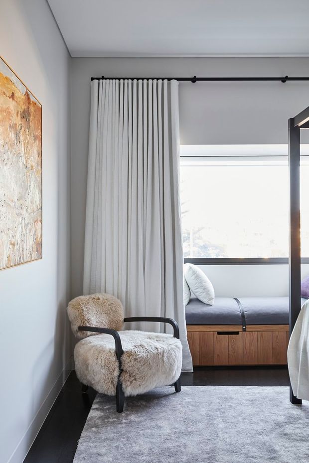 A softly lit bedroom corner with a cream shearling armchair, pale grey rug, and a large abstract artwork on a light grey wall. Full-height white curtains hang from a black ceiling-mounted track beside a built-in window seat with grey upholstery and warm timber cabinetry below. A portion of the bed is visible at right, reinforcing the bedroom setting. The look is refined, contemporary, and textural.
