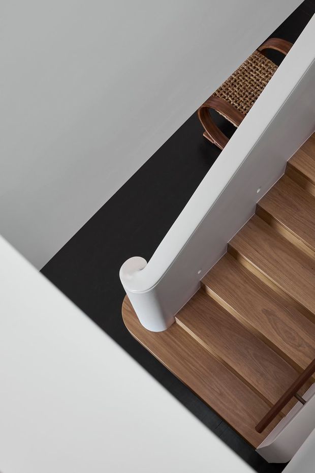 Close-up architectural detail of a modern staircase viewed from above. The stair treads and landing are warm timber, contrasted with a white sculptural balustrade and handrail, black flooring or stairwell void, and a woven timber-and-cane railing detail in the background.