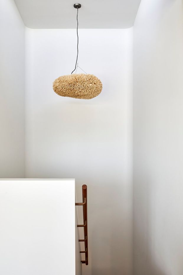 A minimalist white stairwell or landing with tall plain plastered walls and a softly rounded ceiling edge. A sculptural natural-fibre pendant light hangs centrally, while a partial white balustrade and dark timber handrail are visible at the lower left.