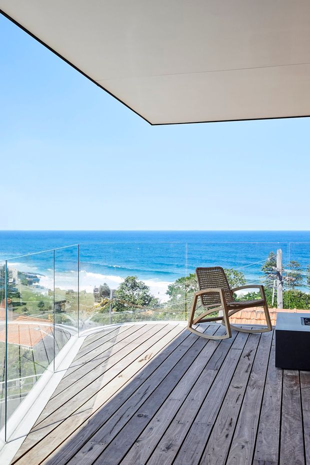 A bright coastal outdoor terrace overlooking the ocean, with weathered timber decking, frameless glass balustrades, a woven timber rocking chair, and a large flat overhead roof or soffit. The view includes waves, beachfront, and nearby coastal homes.