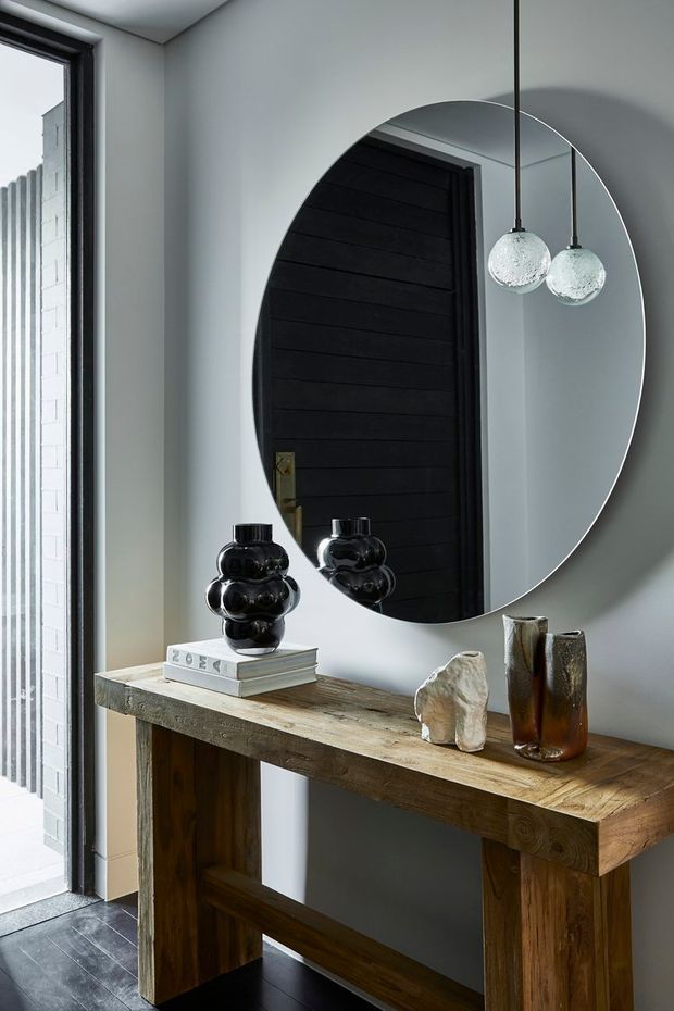 A refined entryway with a rustic timber console table against a pale grey wall, topped with books, black sculptural vessels, ceramic objects and tall brown glass vases. A large oval mirror reflects a dark horizontal timber door and two spherical pendant lights. The space combines natural timber, dark accents and contemporary decorative elements, with a narrow glazed opening at the side.