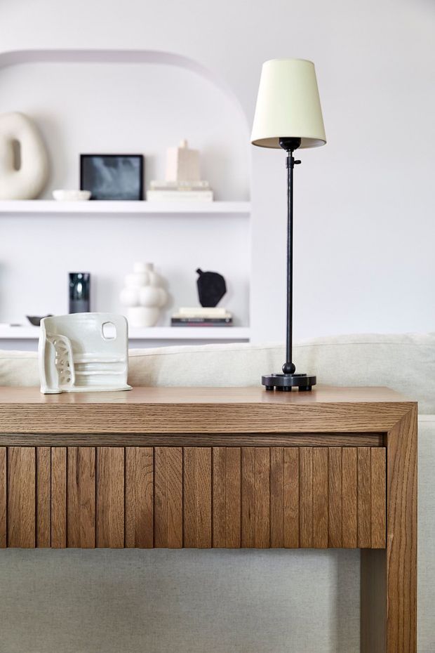 A softly styled living room vignette featuring a warm timber console with vertically fluted cabinet doors, a pale upholstered sofa, and a slim black floor lamp with a cream shade. Built-in white shelving in the background displays neutral decorative objects, framed artwork, books, and ceramics. The overall composition is calm, minimalist, and contemporary with a warm natural palette.