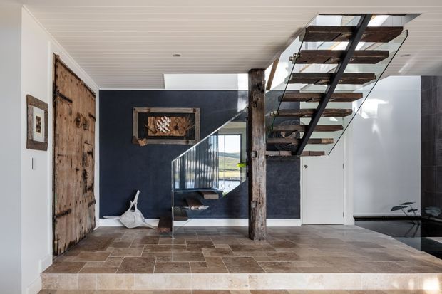 A refined entry hall with a dramatic open-riser timber staircase, exposed rustic timber posts, dark navy feature walls, white panelled ceiling, and large-format stone or tiled flooring. A glass balustrade reveals a stairwell and bright exterior beyond, while rustic timber doors and framed artwork add character.